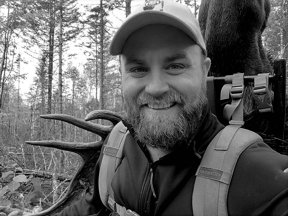 A man with beard and hat standing in the woods.