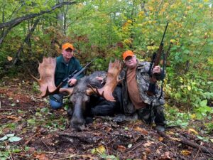 Two men with guns and a moose in the woods.