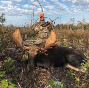 A man in camouflage holding two arrows standing next to a moose.