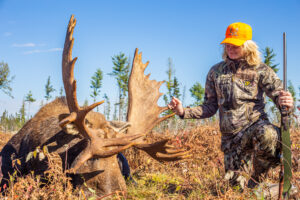 A woman in camouflage and yellow hat standing next to a dead deer.