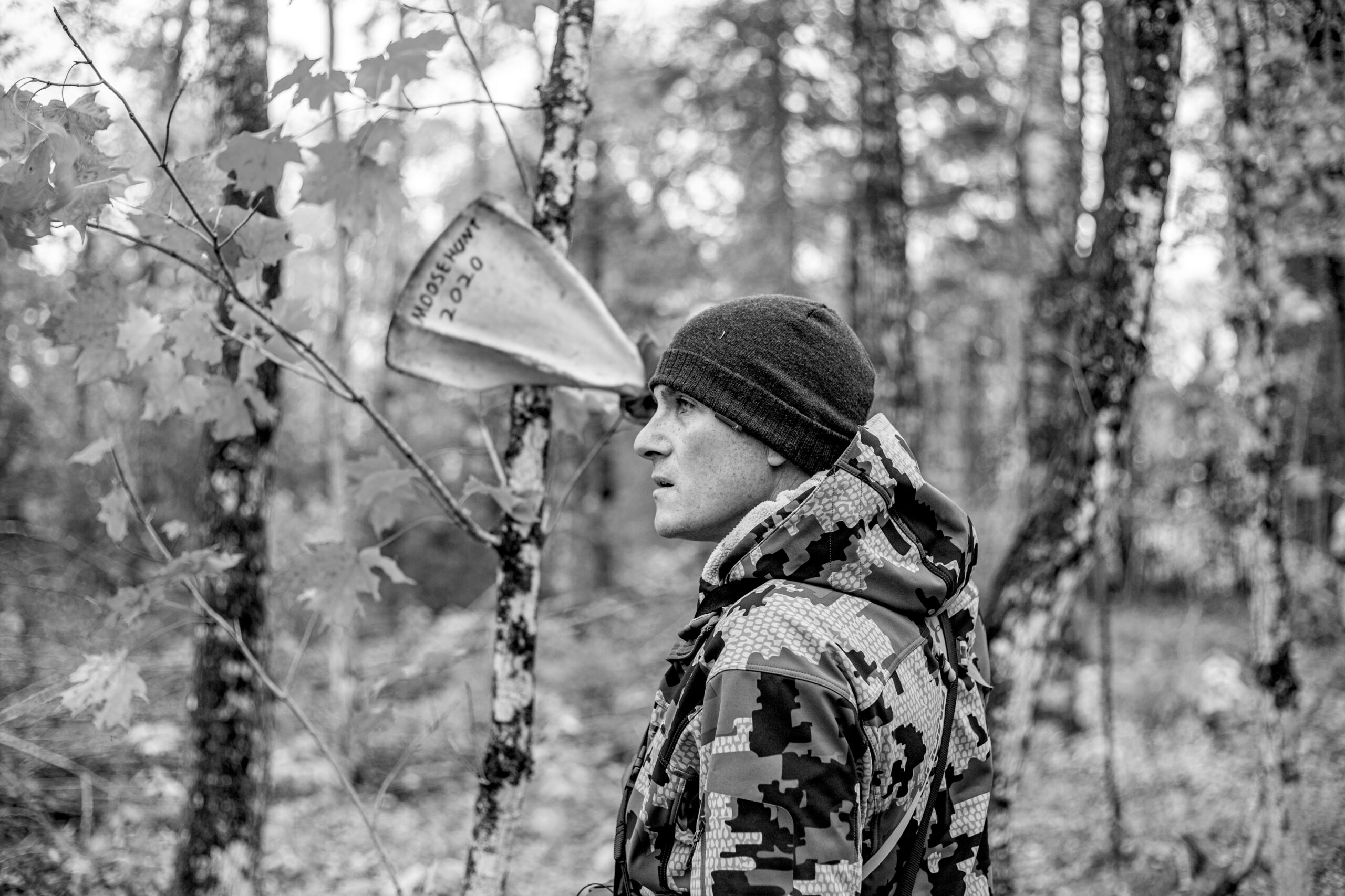 A man in camouflage jacket standing next to trees.